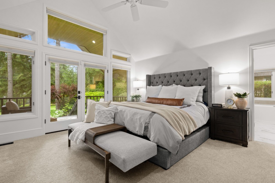 Primary bedroom suite with vaulted ceiling, tufted gray upholstered headboard, French doors to private deck, floor-to-ceiling windows with wooded views, and en-suite bathroom in Canterwood