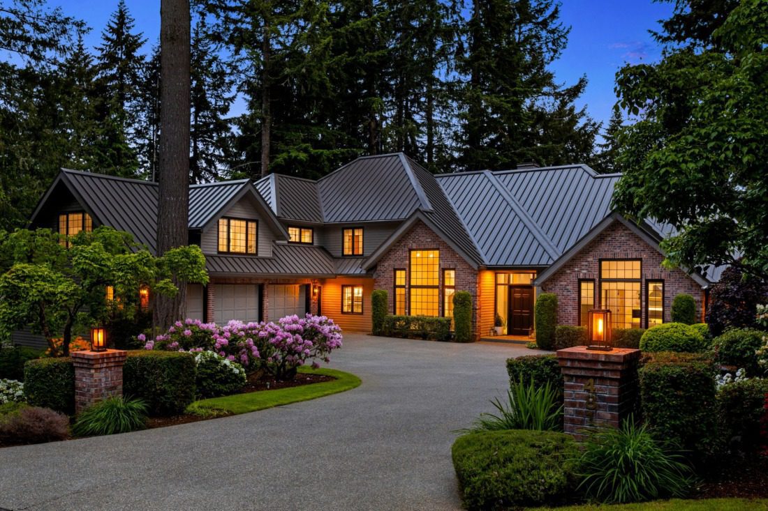 Luxury estate home in Canterwood Golf & Country Club featuring a dramatic brick exterior, metal roof, circular driveway, manicured landscaping, and warm interior lighting surrounded by towering evergreen trees in Gig Harbor, Washington.