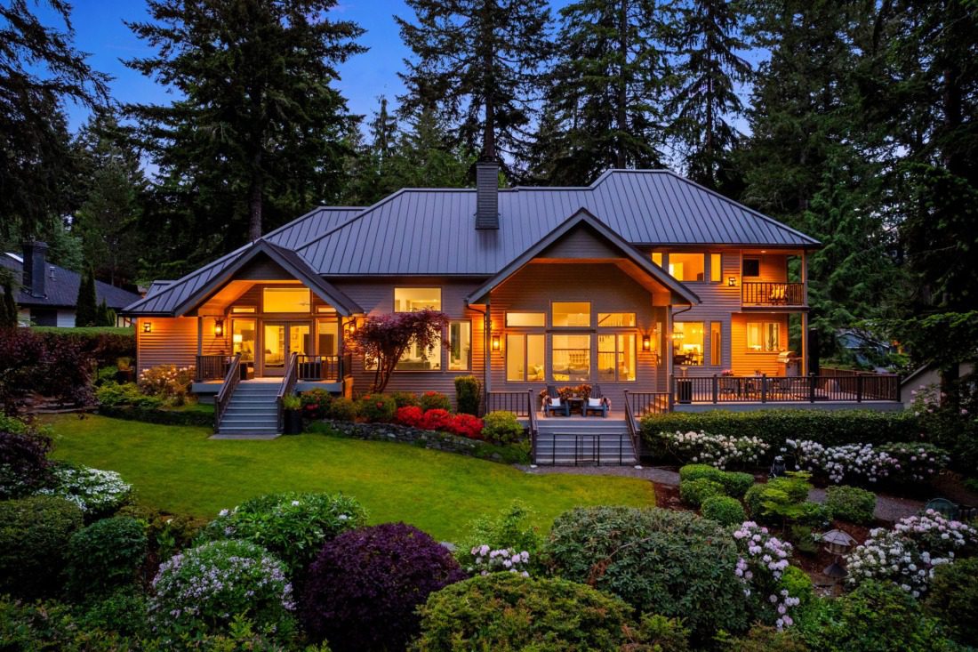 Pacific Northwest luxury home in Canterwood exterior at dusk with standing seam metal roof, wraparound deck, floor-to-ceiling windows, manicured gardens with rhododendrons, and towering evergreen trees